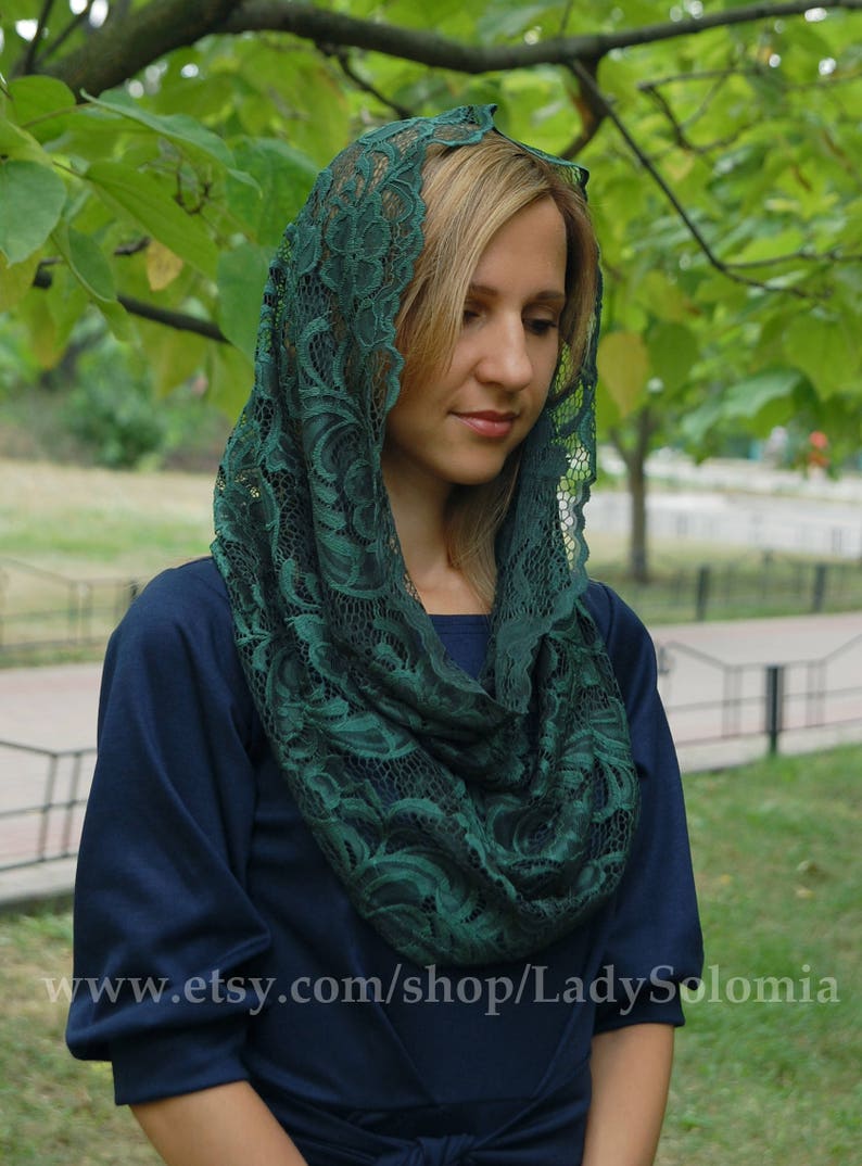May include: A woman wearing a dark green lace shawl over a dark blue dress. The shawl is draped over her head and shoulders, and the lace pattern is intricate and detailed.