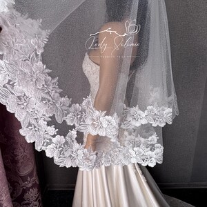May include: A white wedding veil featuring a shimmering, glitter-infused tulle fabric and a wide, scalloped border adorned with intricate white floral embroidery. The veil displays the text Lady Solomia Wedding Veils in white script.