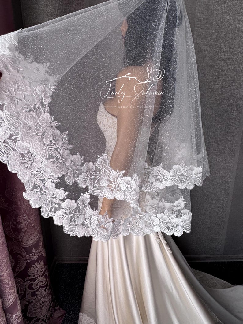 May include: A white wedding veil featuring a shimmering, glitter-infused tulle fabric and a wide, scalloped border adorned with intricate white floral embroidery. The veil displays the text Lady Solomia Wedding Veils in white script.