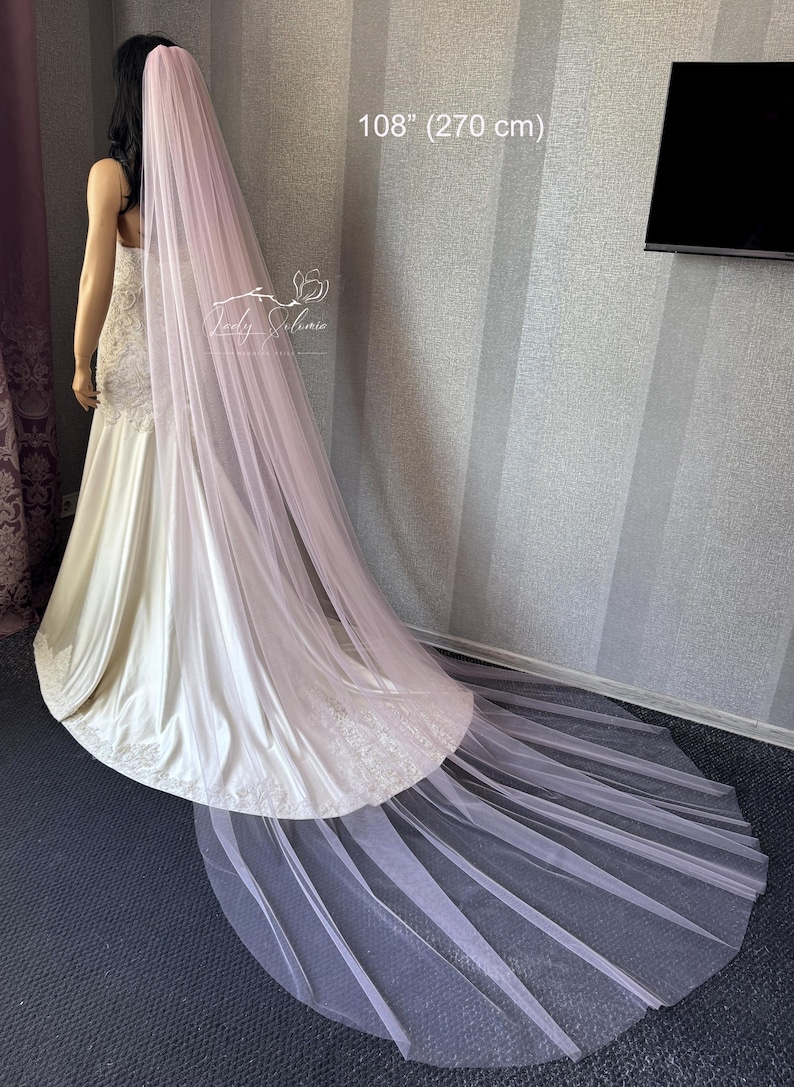May include: A long, pale pink tulle wedding veil measuring 108 inches in length. The veil is draped over a mannequin wearing a white satin gown with lace detailing at the hem. The text 108" (270 cm) is visible in the upper center of the image.
