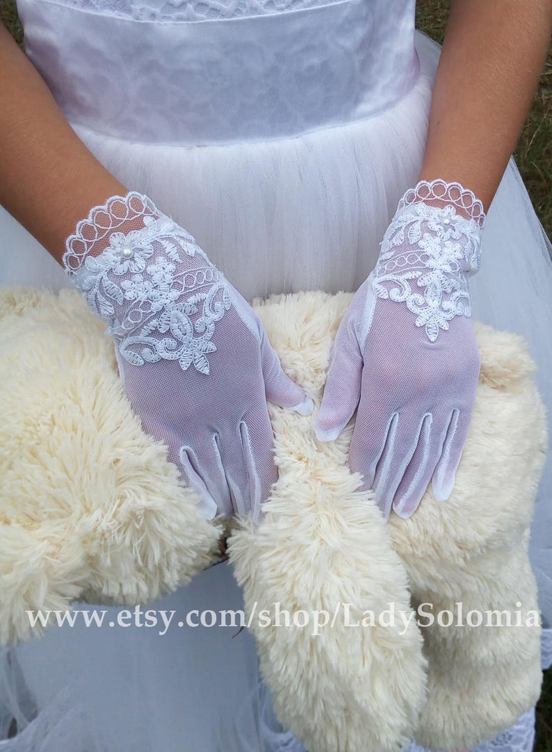 Puede incluir: Un par de guantes blancos hasta la mu&ntilde;eca hechos de tela de malla transparente, con intrincados bordados florales blancos y un borde de encaje festoneado en el pu&ntilde;o. Cada guante tiene una peque&ntilde;a perla centrada en el dorso de la mano.