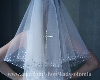 May include: A white two-tier tulle veil featuring a central cross motif made of clear rhinestones and a border embellished with scattered sequins and crystals. The veil is attached to a white satin bow at the base. Text reads www.etsy.com/shop/Ladysolomia.