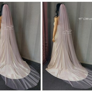 May include: A long, floor-length, pale pink bridal veil made of sheer, lightweight tulle fabric. The veil drapes down the back, extending to a length of 95 inches. It is worn over a cream-colored satin gown with a train that pools on the floor.