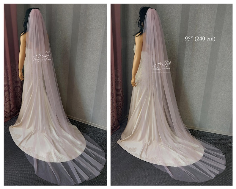 May include: A long, floor-length, pale pink bridal veil made of sheer, lightweight tulle fabric. The veil drapes down the back, extending to a length of 95 inches. It is worn over a cream-colored satin gown with a train that pools on the floor.