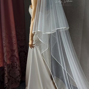 May include: A white bridal veil with a single layer of tulle and a satin edge. The veil is decorated with small, sparkly dots.