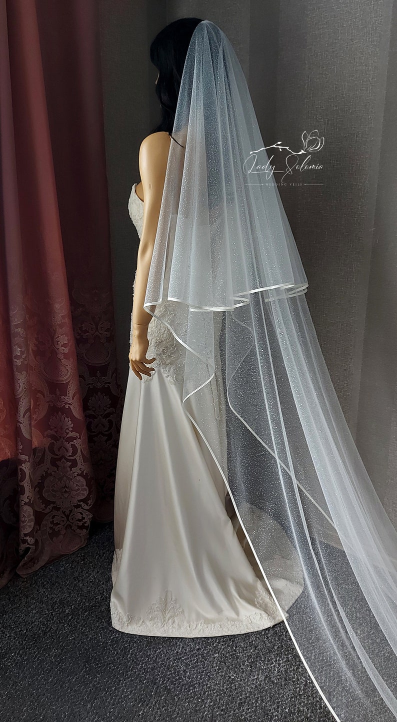 May include: A white bridal veil with a single layer of tulle and a satin edge. The veil is decorated with small, sparkly dots.