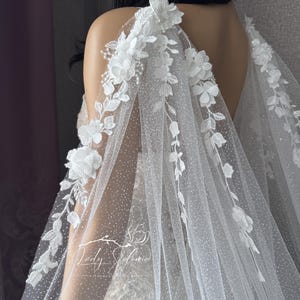 May include: A white bridal cape with floral embellishments. The cape features delicate white flowers and leaves, with a sheer, dotted fabric. The design includes the text "Lady Solomia".