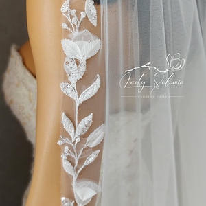 May include: A white bridal veil with a delicate floral design featuring white flowers and leaves with silver embellishments. The veil is made of soft tulle and has a single layer. The text "Lady Solomia Wedding Veils" is visible in the bottom right corner of the image.