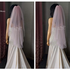 May include: A pale pink, two-tier tulle wedding veil measuring 36 inches in length. The veil is draped over a mannequin wearing a white gown with lace detailing. A logo with the text Lady Silence is visible on the sheer fabric of the veil.