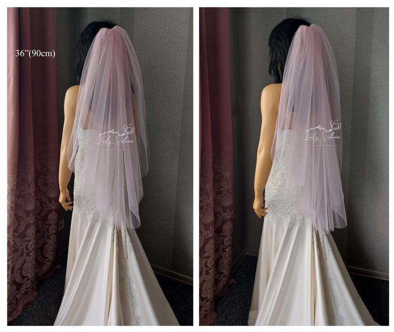 May include: A pale pink, two-tier tulle wedding veil measuring 36 inches in length. The veil is draped over a mannequin wearing a white gown with lace detailing. A logo with the text Lady Silence is visible on the sheer fabric of the veil.