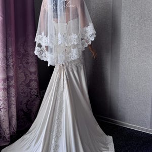 May include: A floor-length ivory wedding dress with a lace-up back and a long train featuring intricate white floral embroidery. A sheer, two-tiered tulle veil with matching white lace trim rests over the shoulders and back, displaying the Lady Solomia logo.