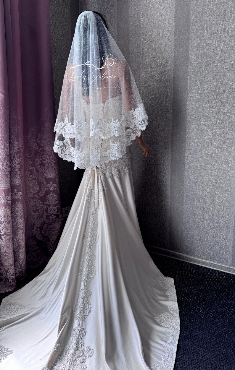 May include: A floor-length ivory wedding dress with a lace-up back and a long train featuring intricate white floral embroidery. A sheer, two-tiered tulle veil with matching white lace trim rests over the shoulders and back, displaying the Lady Solomia logo.