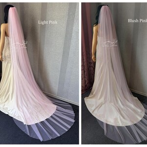 May include: A long, floor-length wedding veil in light pink and blush pink, draped over a mannequin wearing a white gown. The sheer, flowing tulle fabric extends from the head down to the floor, creating a soft, layered effect behind the dress.