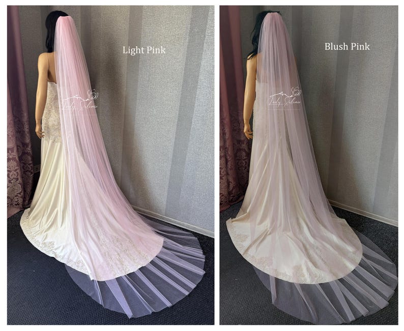 May include: A long, floor-length wedding veil in light pink and blush pink, draped over a mannequin wearing a white gown. The sheer, flowing tulle fabric extends from the head down to the floor, creating a soft, layered effect behind the dress.