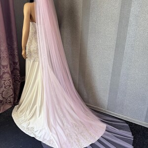 May include: A long, light pink tulle wedding veil draped over a white gown with intricate embroidery along the hem. The veil is sheer, floor-length, and extends beyond the train of the dress. The text Light Pink is visible in the upper right.