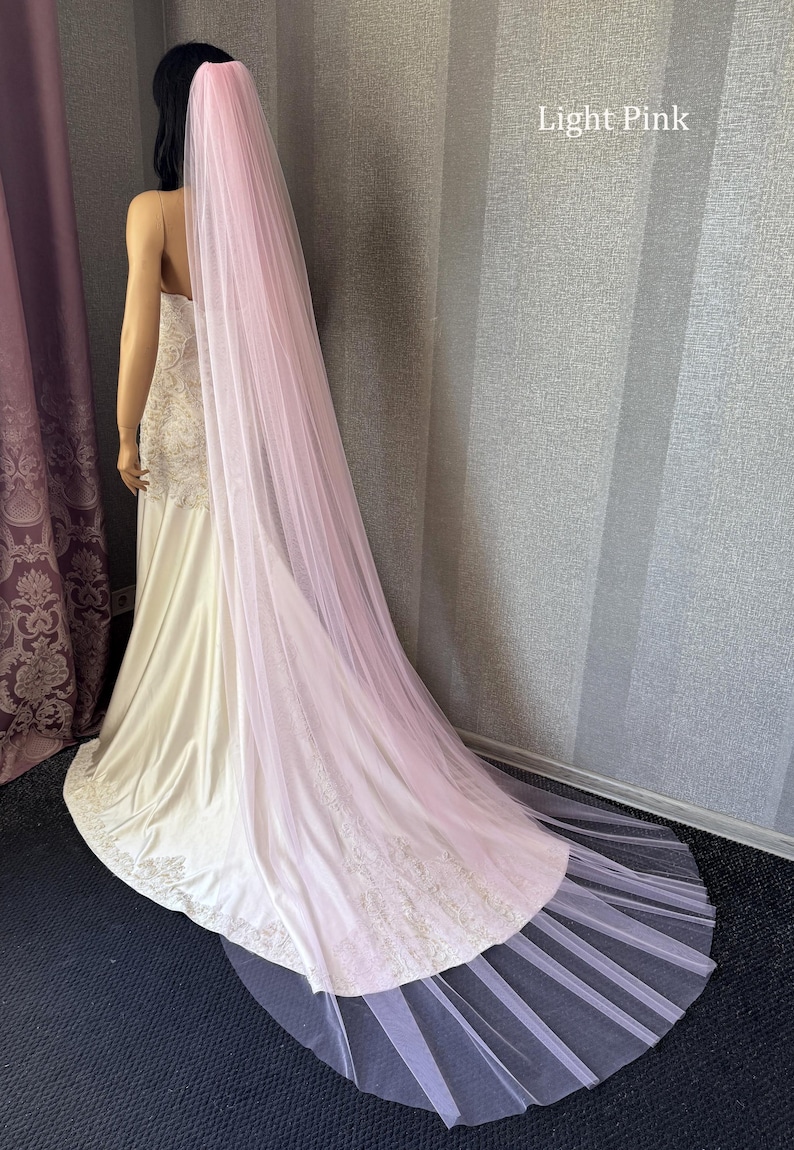May include: A long, light pink tulle wedding veil draped over a white gown with intricate embroidery along the hem. The veil is sheer, floor-length, and extends beyond the train of the dress. The text Light Pink is visible in the upper right.
