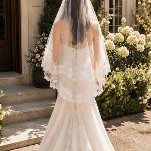 May include: A white wedding veil featuring a delicate lace trim along the edges, layered over a floor-length ivory gown with a fitted bodice and a flared skirt. The veil is sheer and drapes down the back, showcasing intricate floral embroidery.