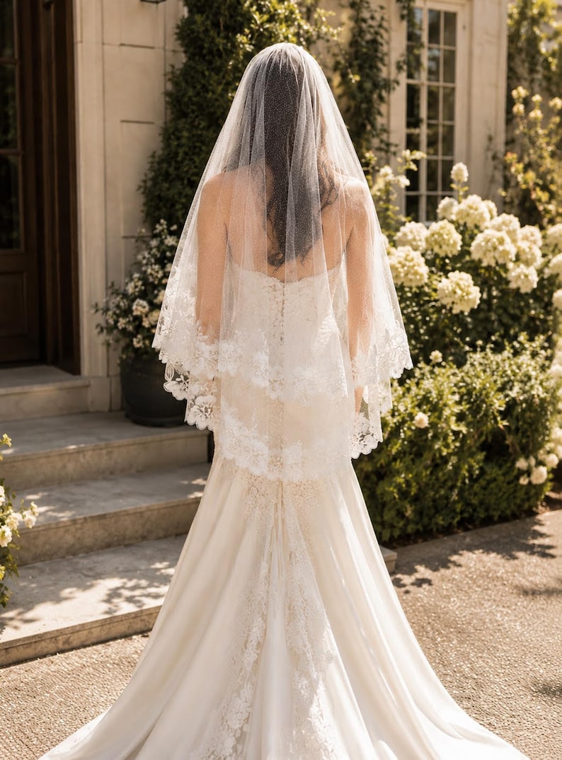 May include: A white wedding veil featuring a delicate lace trim along the edges, layered over a floor-length ivory gown with a fitted bodice and a flared skirt. The veil is sheer and drapes down the back, showcasing intricate floral embroidery.