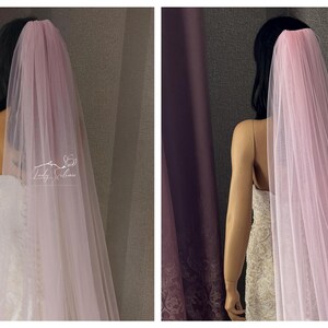 May include: A long, sheer, pale pink tulle veil draped over the back of a mannequin. The veil features a gathered top section and flows downward in soft, translucent layers. A small white logo text is visible on the left side of the fabric.