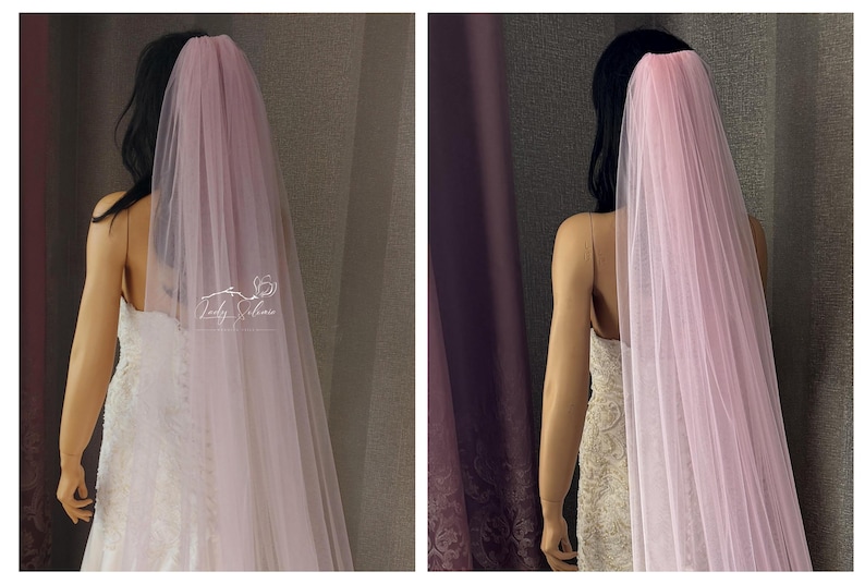 May include: A long, sheer, pale pink tulle veil draped over the back of a mannequin. The veil features a gathered top section and flows downward in soft, translucent layers. A small white logo text is visible on the left side of the fabric.