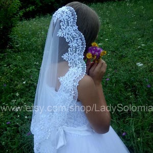 Puede incluir: Un velo de novia blanco con intrincados bordados de encaje floral en los bordes, acentuado con peque&ntilde;as lentejuelas transparentes. El velo est&aacute; sujeto a un vestido blanco con un lazo de cinta de sat&eacute;n en la cintura, la tela es de tul ligero y transparente.
