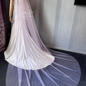 May include: A long, sheer, pale pink bridal veil measuring 108 inches in length. The veil is draped over a white satin gown with lace detailing at the hem. The fabric is lightweight and extends across the floor in a circular train.