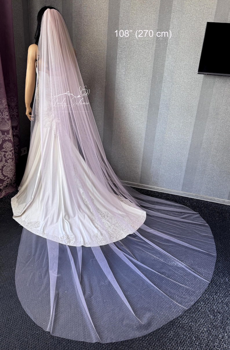 May include: A long, sheer, pale pink bridal veil measuring 108 inches in length. The veil is draped over a white satin gown with lace detailing at the hem. The fabric is lightweight and extends across the floor in a circular train.
