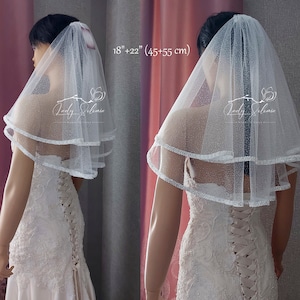 Puede incluir: Un velo de novia blanco de dos niveles hecho de tul brillante con un delicado borde de cuentas. El velo tiene dos capas que miden 45 cm y 55 cm de largo. El texto Lady Solomia es visible en la tela. Se lleva sobre un vestido de encaje.