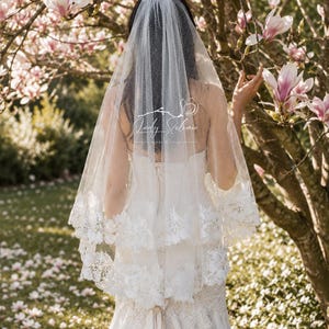 May include: A two-tier white bridal veil featuring a subtle glitter finish and intricate floral lace trim along the edges. The veil is layered over a white gown with lace detailing. A logo with the text Lady Solimia is visible on the veil.