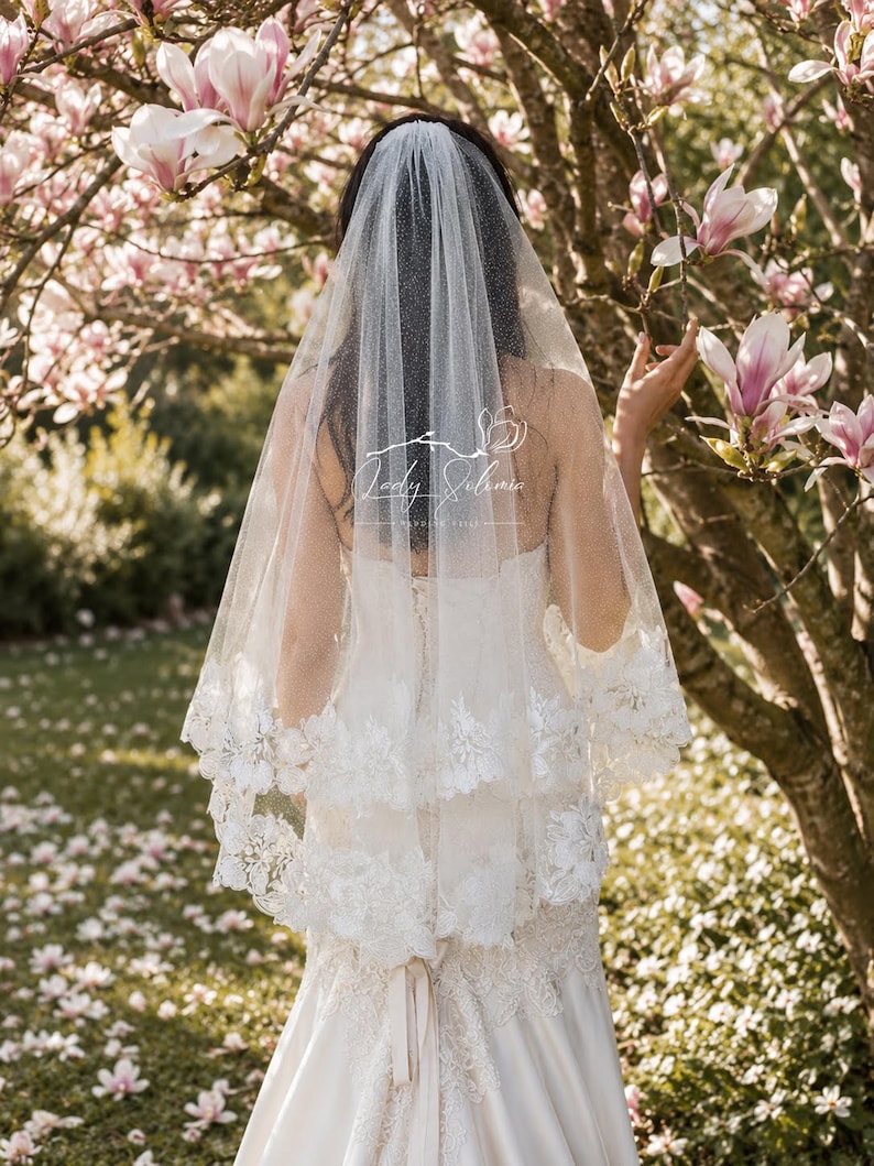 May include: A two-tier white bridal veil featuring a subtle glitter finish and intricate floral lace trim along the edges. The veil is layered over a white gown with lace detailing. A logo with the text Lady Solimia is visible on the veil.