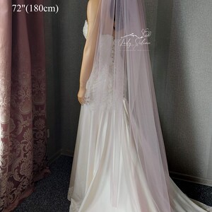 May include: A long, sheer, dusty rose-colored bridal veil made of soft tulle fabric. The veil measures 72 inches in length and is draped over a white satin wedding dress with lace detailing along the hemline and bodice. The text 72"(180cm) is visible.