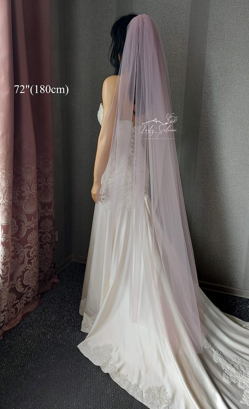 May include: A long, sheer, dusty rose-colored bridal veil made of soft tulle fabric. The veil measures 72 inches in length and is draped over a white satin wedding dress with lace detailing along the hemline and bodice. The text 72"(180cm) is visible.