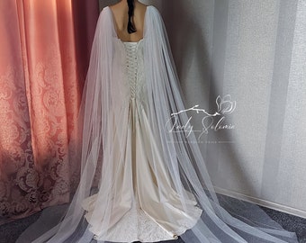 May include: A floor-length ivory gown with a lace-up corset back and a long, sheer, white tulle cape featuring subtle glitter accents. The garment is displayed from the back, showcasing the flowing fabric and the Lady Solomia logo text.