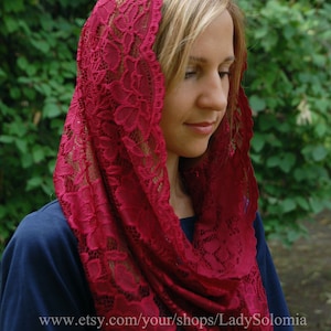 May include: A red lace head covering with a floral pattern. The lace is delicate and intricate. The head covering is worn by a person with blonde hair.