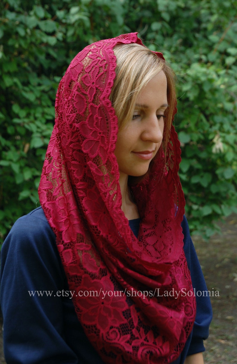 May include: A red lace head covering with a floral pattern. The lace is delicate and intricate. The head covering is worn by a person with blonde hair.