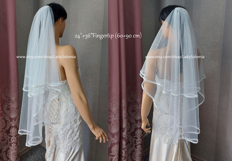 Puede incluir: Un velo de novia de tul blanco de dos niveles con borde de cinta de sat&eacute;n, que mide 60 cm y 90 cm de largo. El velo se muestra en un maniqu&iacute; con un vestido de encaje blanco, con el texto 24"+36"Fingertip (60+90 cm) y una URL.