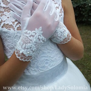 Puede incluir: Un par de guantes de malla blanca semitransparente con intrincados bordados de encaje floral en la mu&ntilde;eca, acentuados con peque&ntilde;as perlas blancas. Los guantes se llevan con un vestido blanco sin mangas con corpi&ntilde;o de encaje y cinturilla de sat&eacute;n.