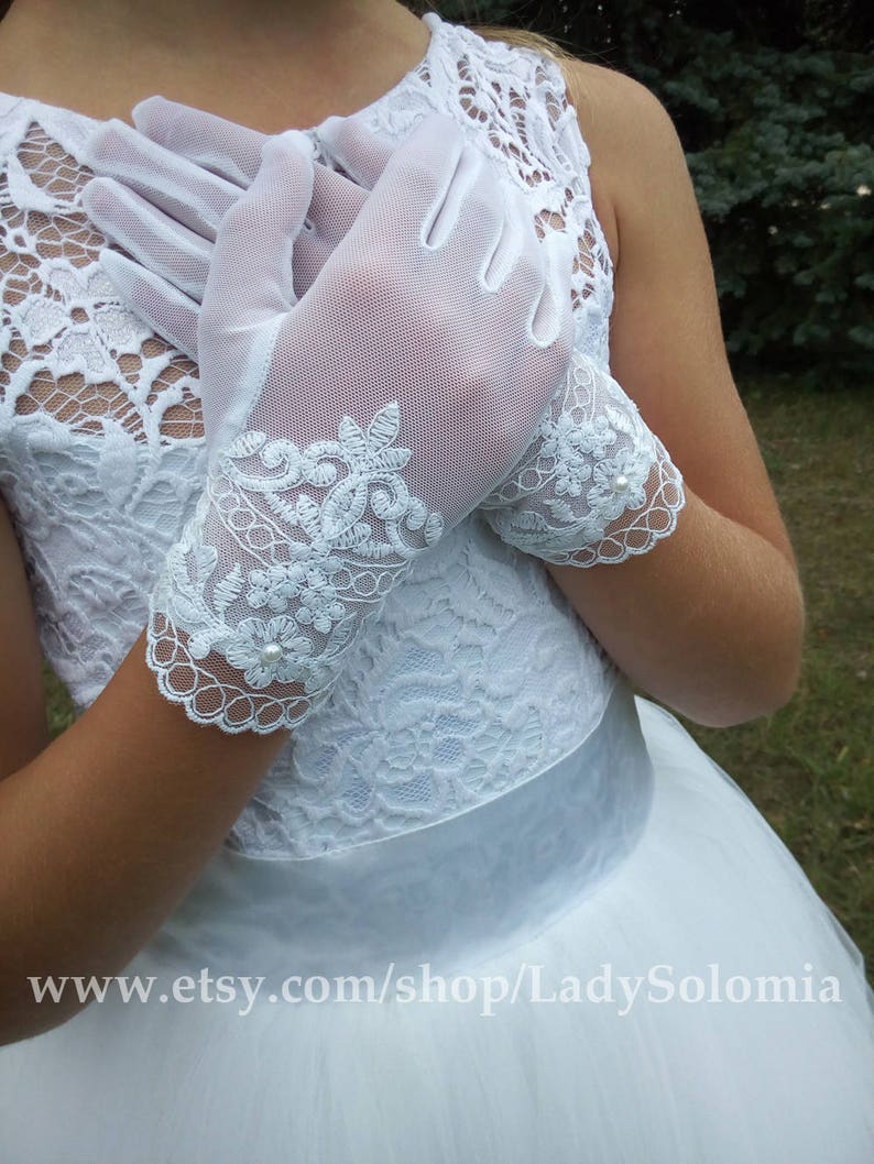 Puede incluir: Un par de guantes de malla blanca semitransparente con intrincados bordados de encaje floral en la mu&ntilde;eca, acentuados con peque&ntilde;as perlas blancas. Los guantes se llevan con un vestido blanco sin mangas con corpi&ntilde;o de encaje y cinturilla de sat&eacute;n.