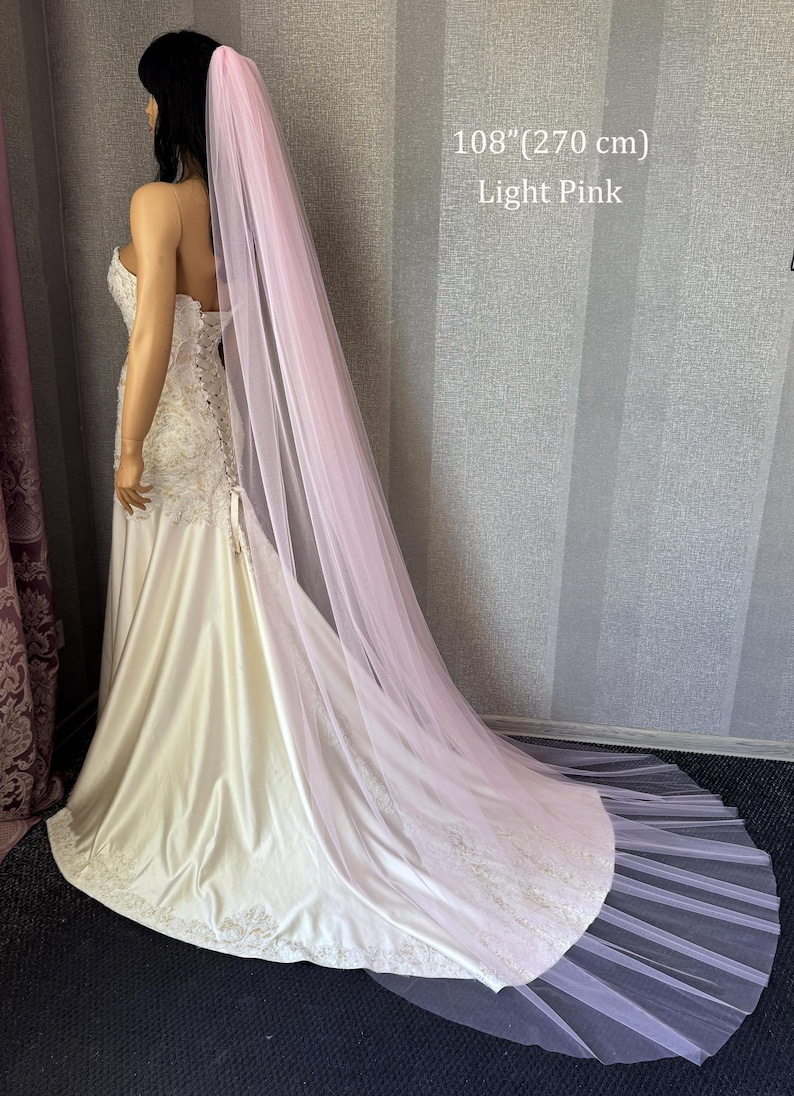 May include: A long, light pink tulle veil measuring 108 inches in length, draped over a strapless, floor-length ivory gown with lace detailing on the bodice and hem. The text 108" (270 cm) Light Pink is visible in the upper right corner.