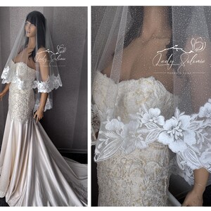May include: A floor-length strapless wedding dress in ivory satin with intricate lace embroidery on the bodice and skirt, paired with a sheer, glitter-dusted veil featuring a wide, scalloped floral lace border. The text Lady Solomia is visible.