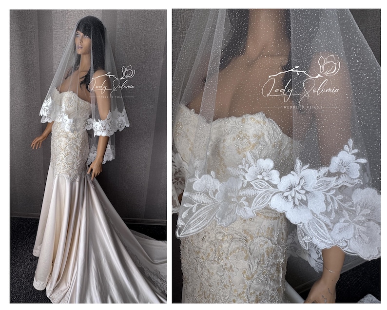 May include: A floor-length strapless wedding dress in ivory satin with intricate lace embroidery on the bodice and skirt, paired with a sheer, glitter-dusted veil featuring a wide, scalloped floral lace border. The text Lady Solomia is visible.