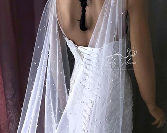 May include: A white floor-length gown featuring a lace bodice with a corset-style lace-up back and sheer tulle cape sleeves adorned with scattered white pearls. The garment includes the text Lady Selonia Wedding Veils printed on the right side.