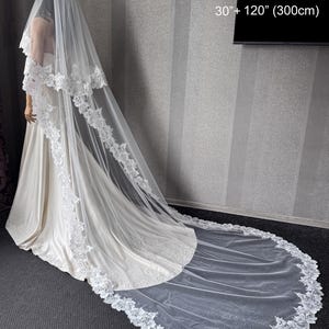 May include: A two-tier white wedding veil featuring intricate floral lace trim along the edges. The veil measures 30 inches and 120 inches in length. It is draped over a gown, extending onto the floor with a long, sheer train.