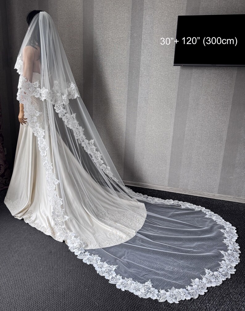 May include: A two-tier white wedding veil featuring intricate floral lace trim along the edges. The veil measures 30 inches and 120 inches in length. It is draped over a gown, extending onto the floor with a long, sheer train.