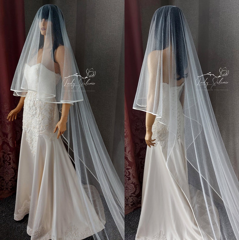 May include: A white tulle wedding veil with a single layer of satin ribbon trim. The veil is shown on a mannequin wearing a white wedding dress.