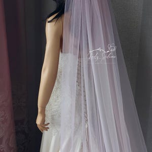 May include: A long, sheer, pale lavender wedding veil made of fine tulle fabric. The veil is draped over a mannequin wearing a white lace and satin gown. A white logo with the text Lady Selinia Wedding Veils is visible on the veil fabric.