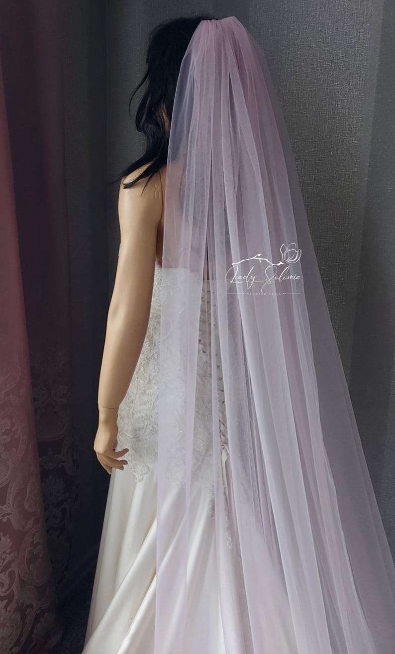 May include: A long, sheer, pale lavender wedding veil made of fine tulle fabric. The veil is draped over a mannequin wearing a white lace and satin gown. A white logo with the text Lady Selinia Wedding Veils is visible on the veil fabric.