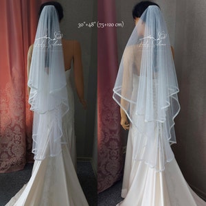Puede incluir: Un velo de novia blanco de dos niveles con acabado brillante y un delicado borde festoneado. El velo mide 75 cm y 120 cm de largo. Se muestra en un maniqu&iacute; que lleva un vestido de sat&eacute;n hasta el suelo con detalles de encaje.