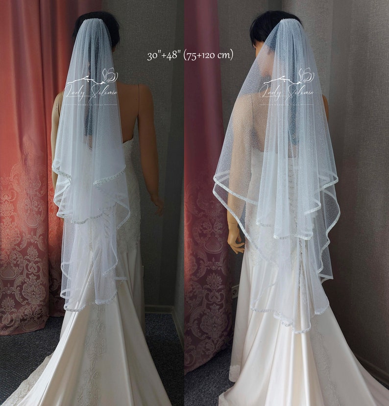 Puede incluir: Un velo de novia blanco de dos niveles con acabado brillante y un delicado borde festoneado. El velo mide 75 cm y 120 cm de largo. Se muestra en un maniqu&iacute; que lleva un vestido de sat&eacute;n hasta el suelo con detalles de encaje.