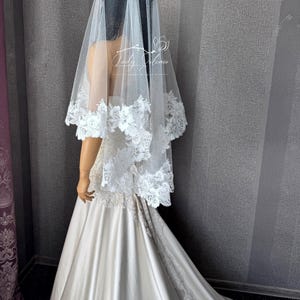 May include: A white wedding veil featuring a shimmering tulle fabric and a wide border of intricate floral lace appliqu&eacute;. The veil is draped over a mannequin wearing a floor-length satin gown with a train. The text Lady Alonia is visible.